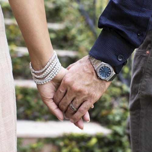 Close up of man and woman holding hands wearing wedding band, watch, and sparkling diamond bracelets